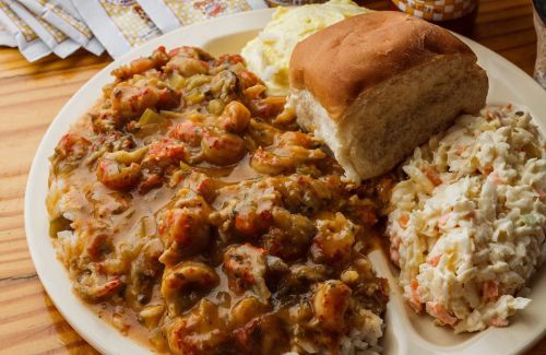 A plate of crawfish étouffée with rice, coleslaw, a roll, and a side of mashed potatoes on a wooden table.
