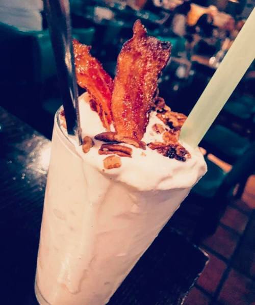 A creamy milkshake topped with crispy bacon and nuts, served in a tall glass with two straws.