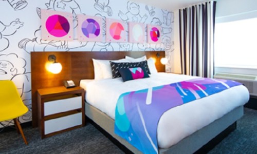 A modern hotel room featuring a large bed with colorful bedding, playful wall art, and stylish furniture.