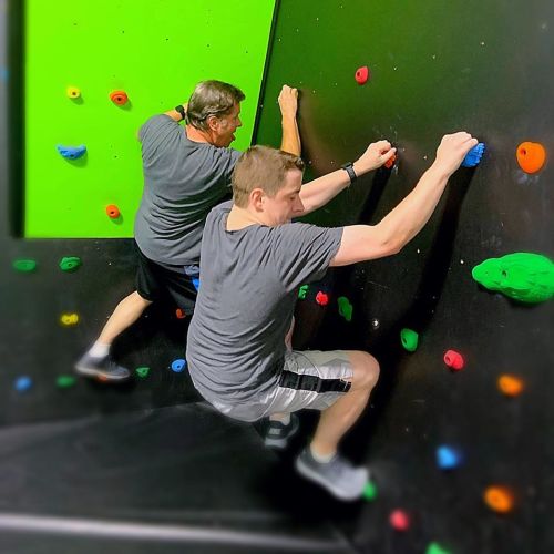 Two men climbing a colorful indoor rock wall, focused on their ascent. Bright green and black background.