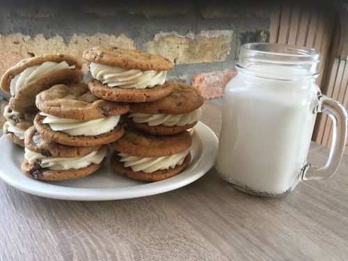A plate of chocolate chip cookie sandwiches filled with frosting, next to a glass of milk on a wooden table.