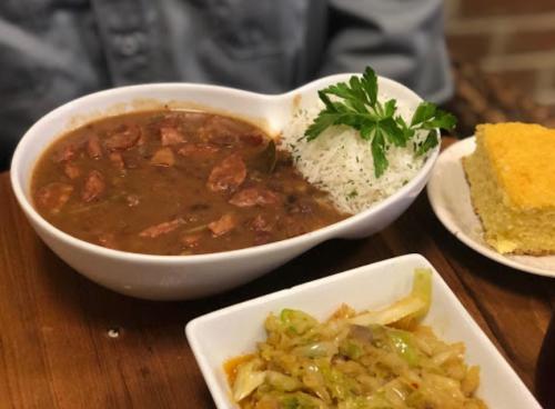 A bowl of stew with sausage, served with rice and parsley, alongside a plate of cabbage and a slice of cornbread.