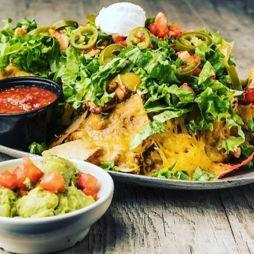 A plate of nachos topped with lettuce, jalapeños, tomatoes, cheese, and a side of guacamole and salsa.