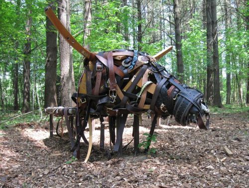 A sculpture of a dog made from various leather straps and materials, set in a wooded area with trees in the background.