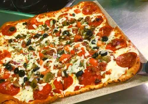 A rectangular pizza topped with pepperoni, olives, tomatoes, green peppers, and drizzled with white sauce.