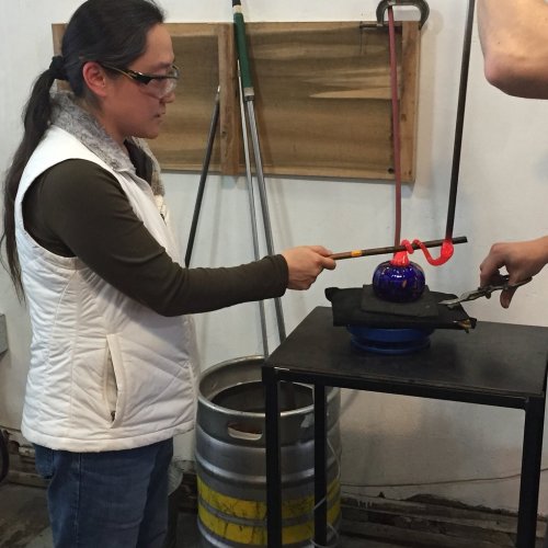 A person in a white vest shapes molten glass with tools at a glassblowing workshop.