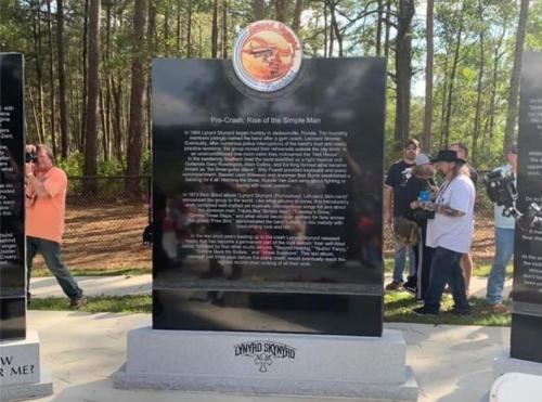 A black monument with text about Lynyrd Skynyrd's history, surrounded by trees and people in the background.