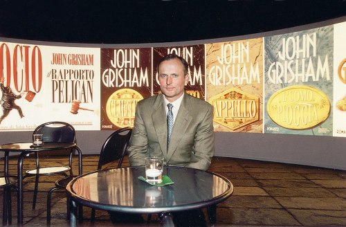 A man in a suit sits at a table in front of large book covers by John Grisham displayed on a curved backdrop.