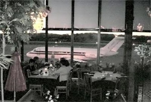 A view from a restaurant window showing a plane parked outside, with diners seated at tables in the foreground.
