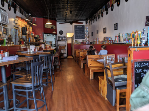 Interior of a cozy restaurant with wooden tables, black chairs, and a bar area, featuring a warm, inviting atmosphere.