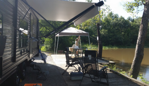 A camper trailer by a serene lake, with a person setting up a table under a canopy and outdoor chairs nearby.
