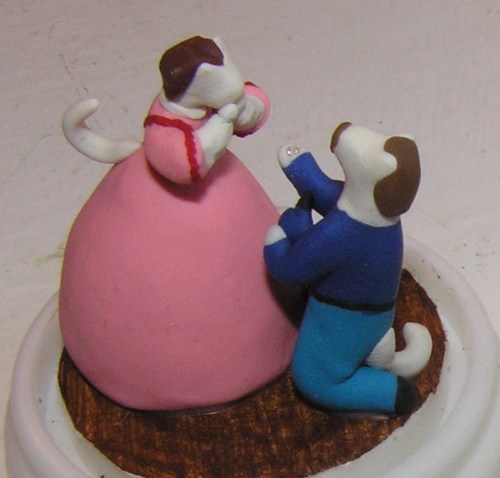 A clay figurine of a dog proposing to another dog in a pink dress, set on a circular base.