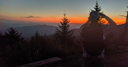 A person with a backpack takes a photo of a vibrant sunset over mountain ridges, surrounded by trees.