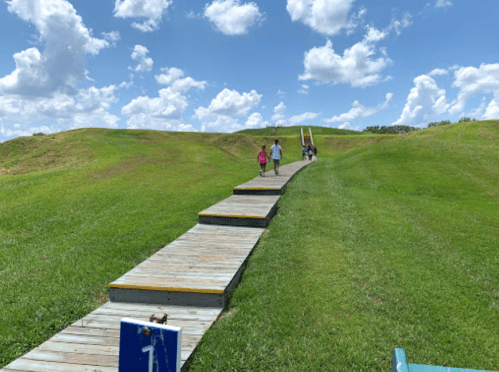 A wooden walkway leads up a grassy hill under a blue sky with fluffy clouds, with people walking towards a slide.