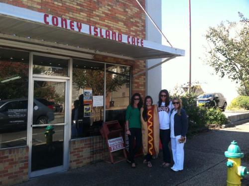 Four women pose outside Coney Island Cafe, one wearing a hot dog costume, with a brick building in the background.