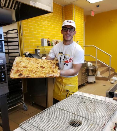 A smiling young man in a white shirt and yellow apron holds a large tray of freshly baked pizza in a bright kitchen.