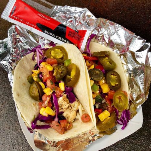 Two tacos filled with grilled chicken, corn, tomatoes, jalapeños, and purple cabbage, with a packet of sour cream on the side.