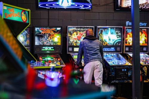 A person in a denim jacket plays pinball at a retro arcade, surrounded by colorful machines featuring various themes.