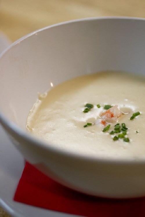 A creamy soup in a white bowl, garnished with shrimp and chopped chives, resting on a red napkin.