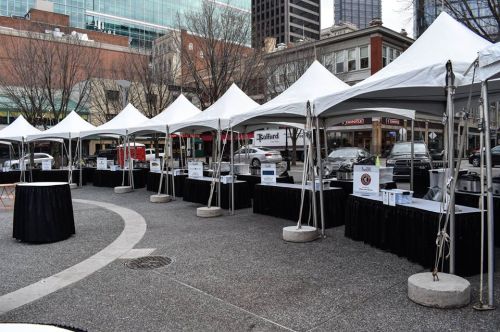 Row of empty vendor tents set up in an urban plaza, with tables and signage, surrounded by buildings and parked cars.
