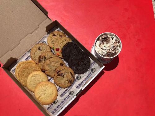 A box of assorted cookies next to a cup of chocolate swirl ice cream on a red surface.
