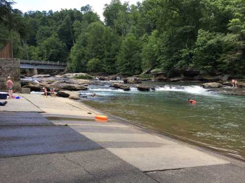 A serene river scene with people enjoying the water, surrounded by lush greenery and a bridge in the background.