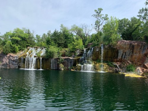 A serene landscape featuring a rocky cliff with two waterfalls cascading into a calm green lake surrounded by trees.