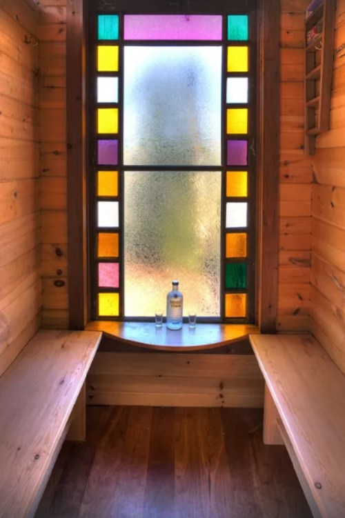 A cozy wooden sauna with a colorful stained glass window and two benches, featuring a bottle and glasses on a ledge.