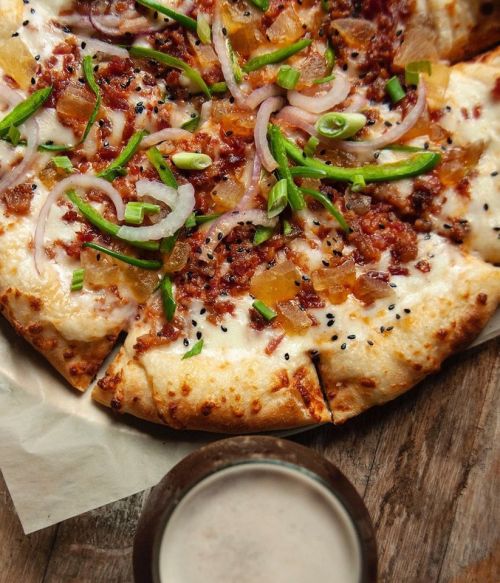 A close-up of a pizza topped with cheese, bacon, onions, and green onions, served with a drink on a wooden table.