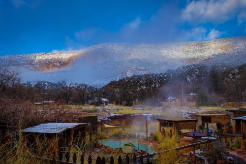 A scenic view of misty hot springs surrounded by mountains, with snow on the peaks and a blue sky above.