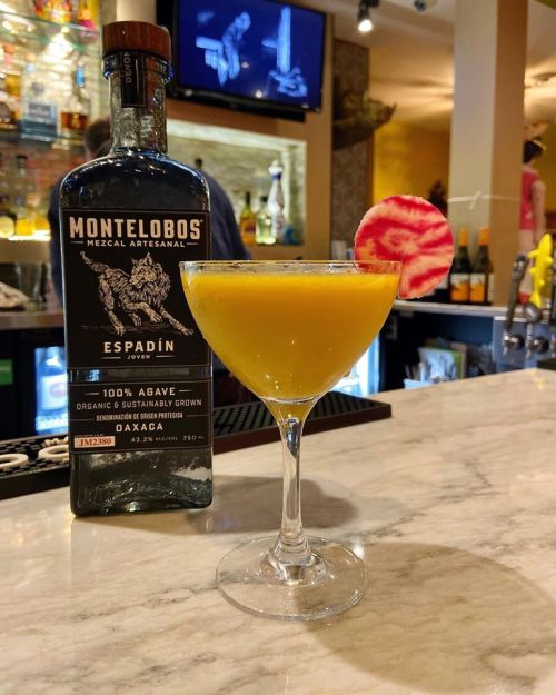 A vibrant yellow cocktail garnished with a pink slice, next to a bottle of Montelobos Mezcal on a marble bar.