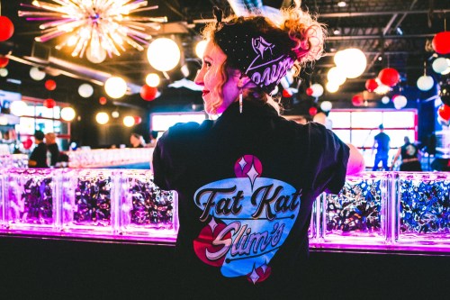 A person with curly hair wearing a black shirt with "Fat Kat Slim's" logo stands at a vibrant bar with colorful decor.