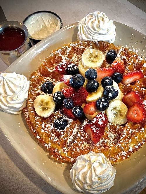 A plate of fluffy pancakes topped with bananas, strawberries, blueberries, whipped cream, and syrup on the side.