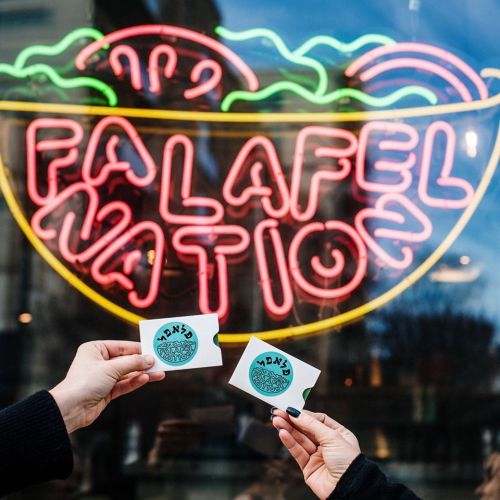 Two hands hold cards in front of a neon sign that reads "Falafel Nation," featuring a colorful falafel graphic.