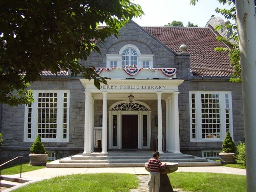 Front view of Derby Public Library, featuring stone architecture, large windows, and patriotic bunting. A child stands nearby.