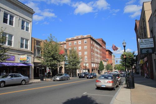 A street view featuring shops, trees, parked cars, and a flag against a blue sky with clouds.