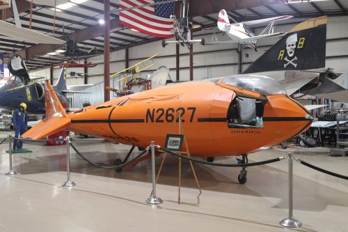 An orange experimental aircraft displayed in a hangar, surrounded by other planes and American flags.