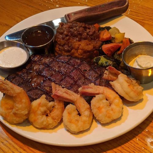 A plate featuring grilled steak, shrimp, baked beans, and mixed vegetables, served with dipping sauces.