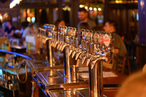 A row of beer taps at a bar, with patrons in the background enjoying drinks and conversation.