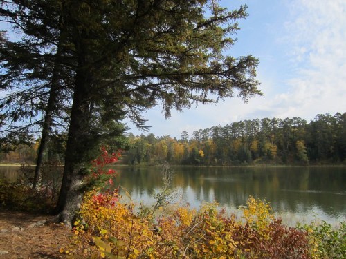 A serene lake surrounded by trees and colorful autumn foliage under a partly cloudy sky.