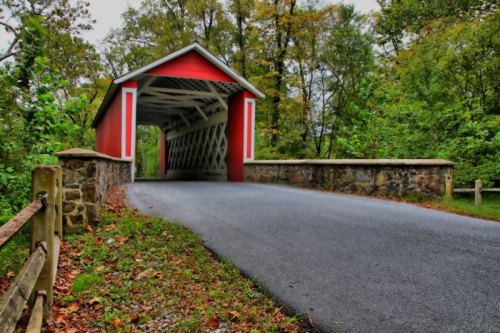 A red covered bridge with a stone wall, surrounded by trees and autumn foliage, leading to a paved road.