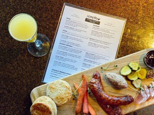 A wooden platter with biscuits, sausage, carrots, pickles, and a menu beside a glass of mimosa on a table.