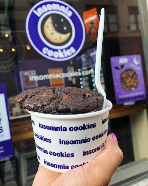 A hand holding a cup of cookies from Insomnia Cookies, with a chocolate cookie on top and a spoon.