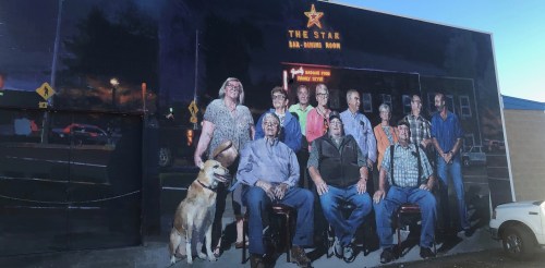 Mural of a group of people and a dog in front of a bar, with a star sign and evening sky in the background.