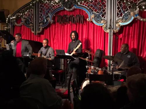 A live band performs on stage at a venue with red curtains, featuring a singer, guitarist, and drummer.