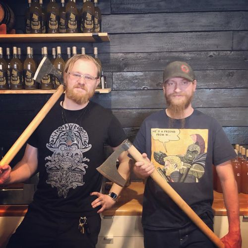 Two bearded men stand in a bar, each holding a large axe, with bottles lined up on a shelf behind them.