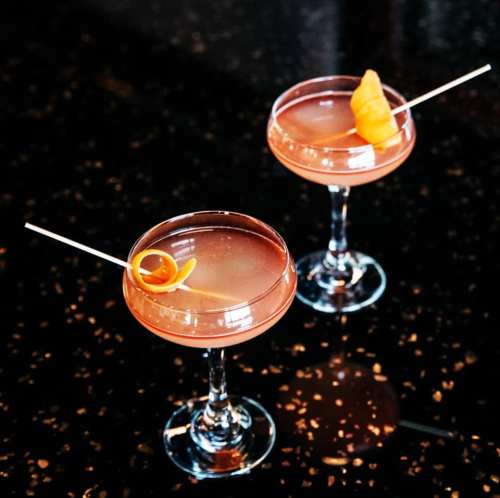 Two elegant cocktails in coupe glasses, garnished with orange twists, on a dark, speckled countertop.
