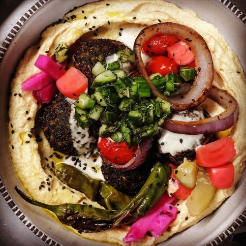A bowl of hummus topped with falafel, fresh vegetables, pickles, and drizzled with sauce, garnished with herbs.