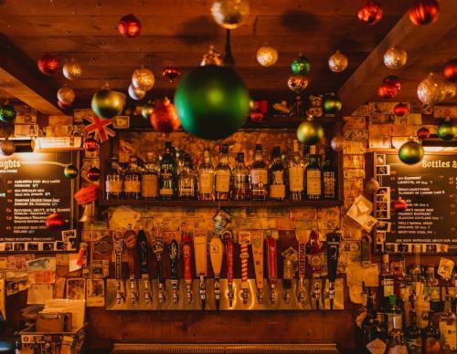 A cozy bar with a wooden interior, decorated with festive ornaments and a variety of liquor bottles and beer taps.