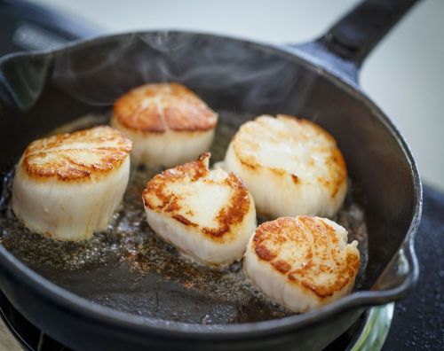 Five perfectly seared scallops sizzling in a cast iron skillet, with steam rising from the hot oil.